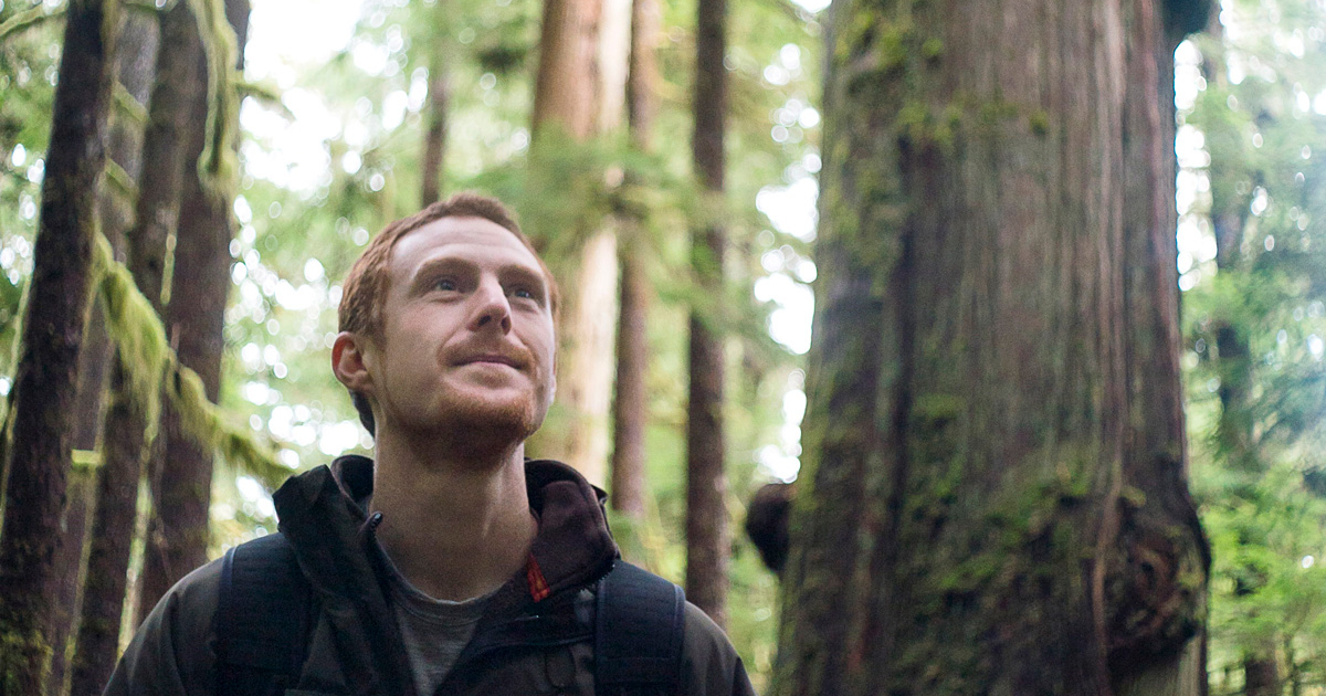 TJ stands in the forefront wearing a black hoodie. Behind him stands a number of old-growth cedars and other ancient trees.