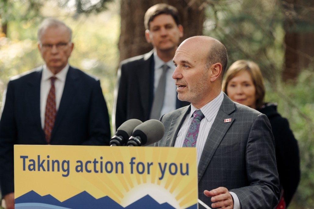 Nathan Cullen stands at a podium with the text, "Taking action for you," while he makes an announcement. Premier Eby and two other government officials stand behind him.