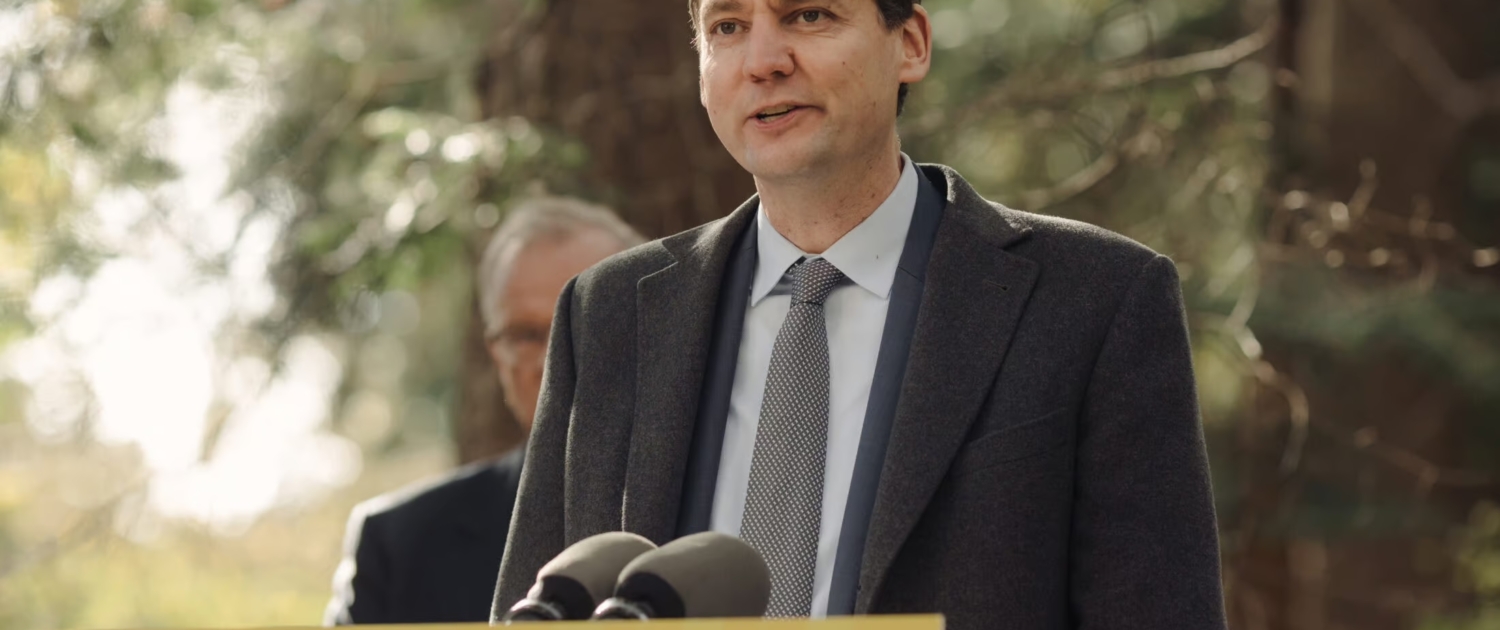 Premier David Eby stands at a yellow podium that reads, "Taking action for you," with trees in the background.